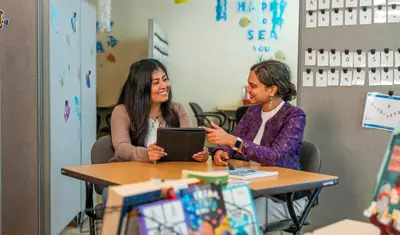 A psychology student and faculty member sit together reviewing material on a tablet, sharing an encouraging and engaged conversation in a classroom decorated with ocean-themed elements and educational tools.