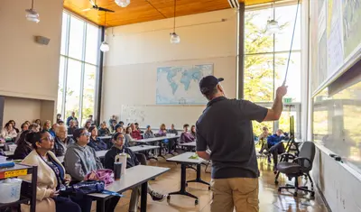 Professor standing up next to a board, lecturing students in a classroom.