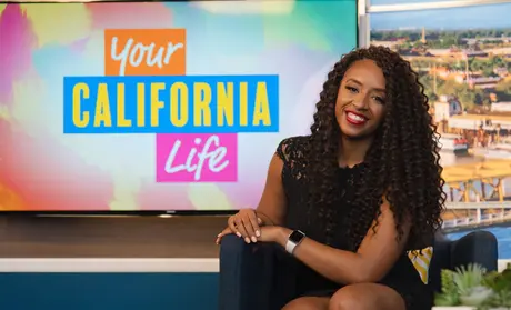 Desiree Sheppard sitting on a couch in front of a TV that shows a text graphic that reads 