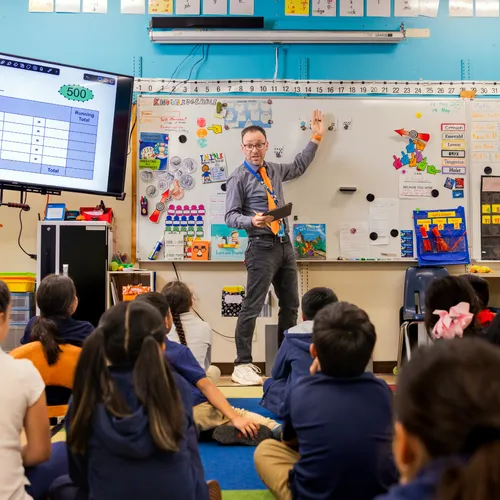 Teacher with a tablet points to the whiteboard while speaking to students seated on the classroom floor