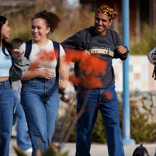 4 students walking and talking, one of them is holding a scooter as they walk past orange flowers