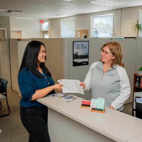 A CSUMB employee with long black hair hands a document to an employee with short blond hair standing behind a reception counter in a bright, simple office.