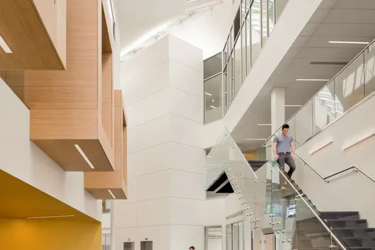 Modern university or office building interior with high ceilings, glass railings, and natural light streaming through skylights. People are walking, sitting, and using the stairs in the open, airy atrium.