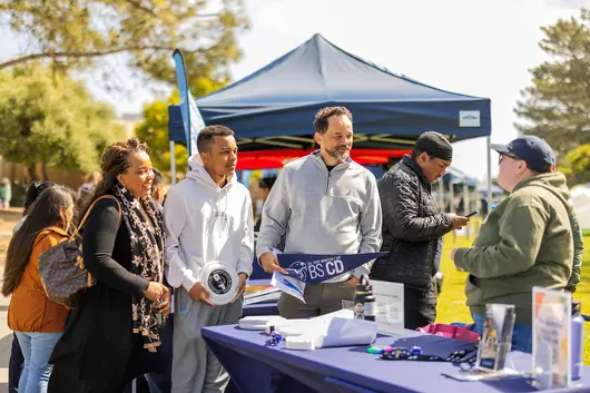 Family of three is outside, smiling and speaking to a staff member at a table