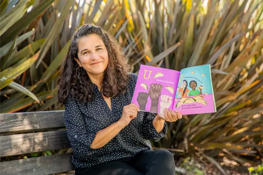 A woman sitting on a bench outdoors smiles while holding open a colorful children’s book featuring illustrations of raised fists and people eating tacos, with text in both English and Spanish.