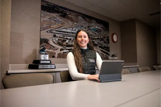 A student sitting at a desk smiling at the camera while she has her computer open on the desk.
