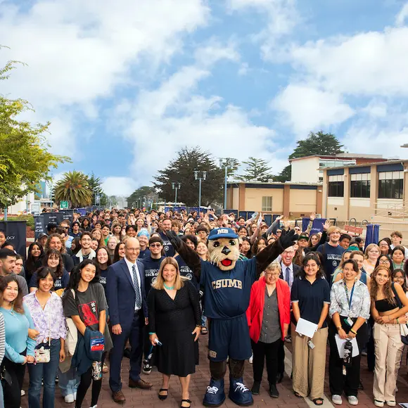 A large group of students, staff and the campus mascot gather outdoors, celebrating community and belonging at Cal State Monterey Bay.