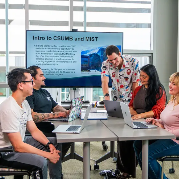 Several students sit around a table in front of a screen that reads 