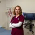 A portrait of a nursing student in a scrub with a stethoscope wrapped around their neck smiling at the camera with a bed and computer in the background