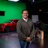 Film crew member posing for photo in front of a green screen and a red car behind him.