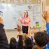 Teacher stands at the whiteboard while students raise their hands in class