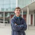 A student standing with his arms crossed and smiling for a portrait in front of a building
