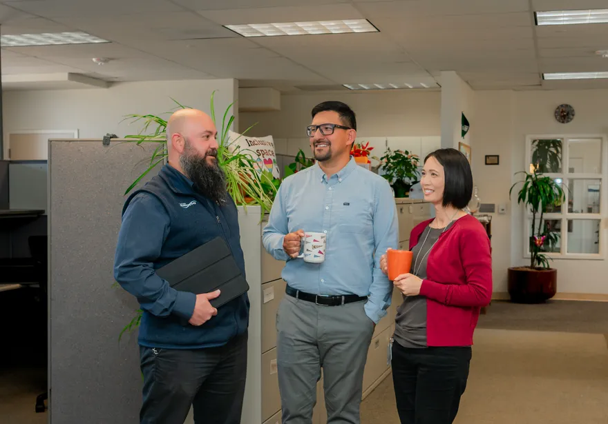 Three CSUMB HR staff members share a friendly conversation near a cubicle partition lined with plants and an “Inclusive Space” sign, smiling and holding coffee mugs and a tablet.