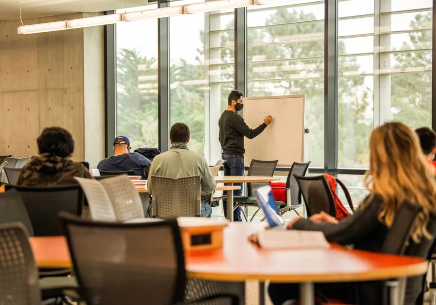 Instructor writing on a whiteboard while students listen in a classroom.