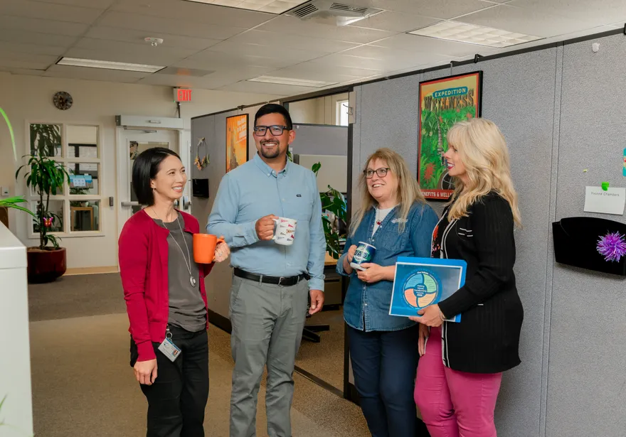 Four CSUMB HR staff members enjoy a casual coffee break together in a collaborative office space, laughing and chatting with colorful mugs and team materials in hand.