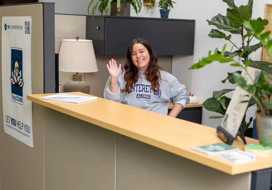 A woman stands behind a desk, smiling warmly and waving hello.