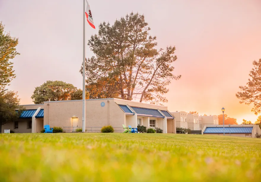 A campus lawn glows at sunset with buildings, trees, and U.S. and a California flag under a sky filled with golden clouds.