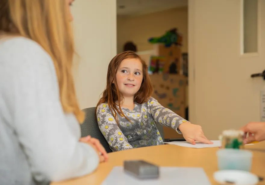 A young girl sitting at a classroom table smiles and points at a piece of paper.