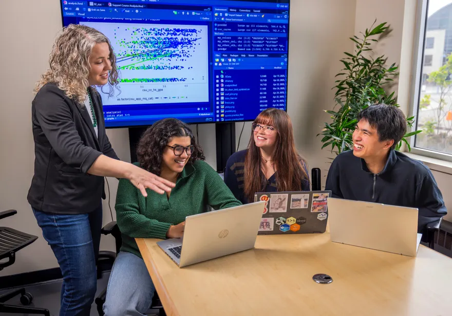 A standing teacher is being watched by two people, while one student looks at a laptop, all of them smiling
