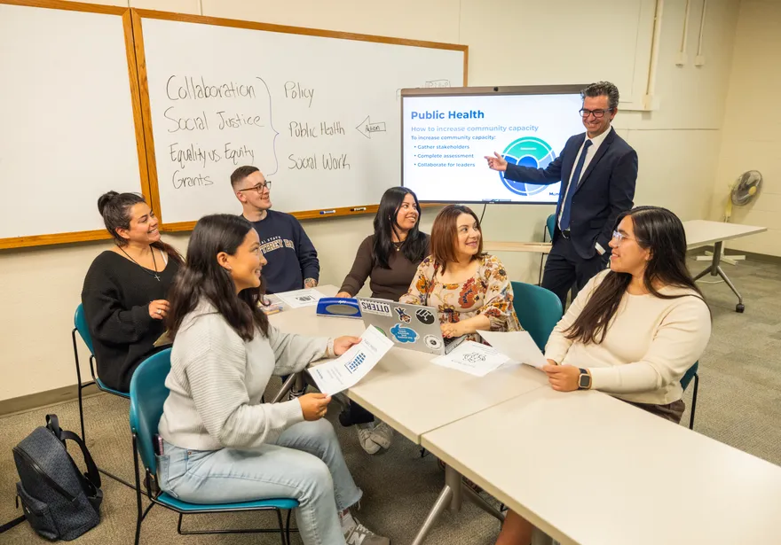 Six students sitting at a table listening and taking notes while a professor stands, teaching them in front of a white board and screen.