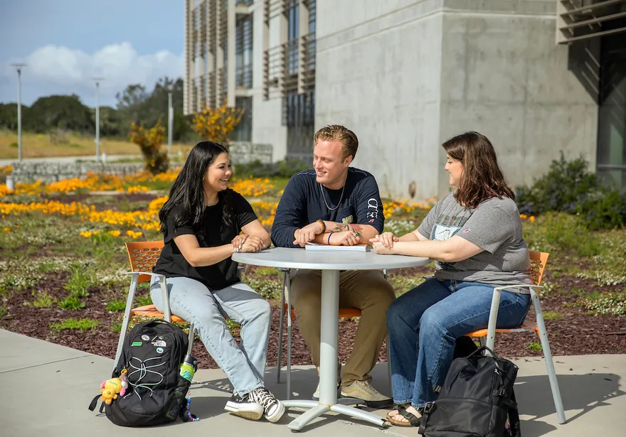 Students sit together outdoors at a campus table, building connections through conversation in a relaxed and supportive setting.