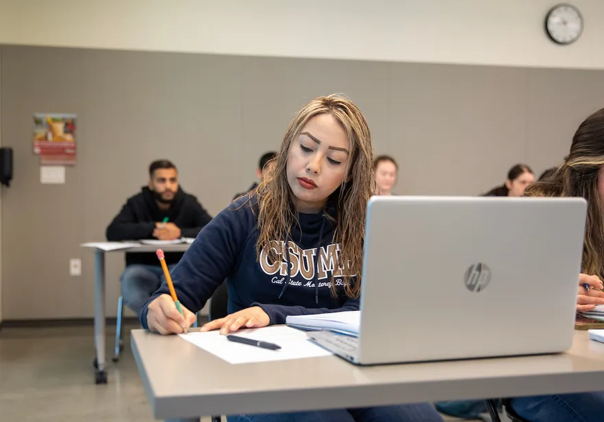 A student takes notes during a classroom session while using a laptop, demonstrating focus and active learning.