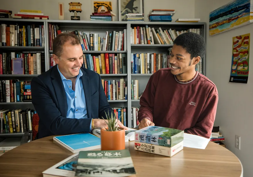A faculty member and a student sit together at a table in an office lined with bookshelves, smiling as they review Japanese-language books and written materials during an academic discussion.