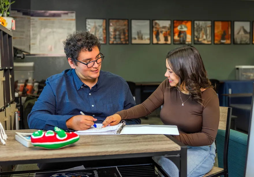 A CSUMB instructor and a student sit together at a table during a one-on-one Spanish language learning session. The instructor writes in a notebook while the student points and smiles, engaging in conversation. Bookshelves and cultural imagery in the background create a supportive and focused learning environment.
