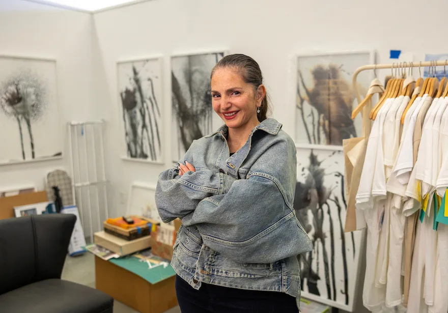 A faculty member stands with arms crossed in an art studio filled with framed artwork and displayed apparel, smiling toward the camera.