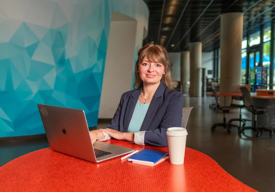 A CSUMB instructional science and technology faculty member sits at a table with a laptop, posing for a professional portrait in a campus learning space.