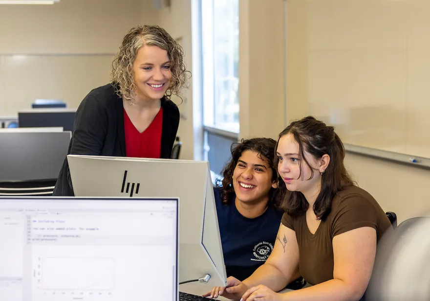 A faculty member works with two students at computer stations in a classroom highlighting mentoring, collaboration and hands-on learning at Cal State Monterey Bay.
