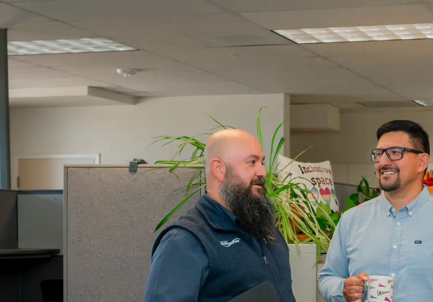 Three ���Ķ���vlog HR staff members share a friendly conversation near a cubicle partition lined with plants and an “Inclusive Space” sign, smiling and holding coffee mugs and a tablet.
