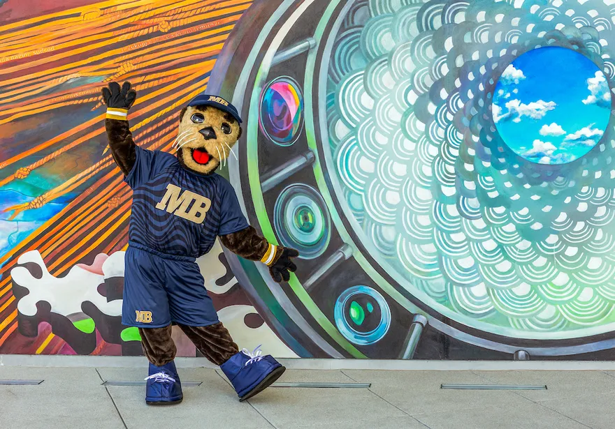 The campus mascot poses in front of a vibrant mural, showcasing creativity and school spirit in an artistic setting.