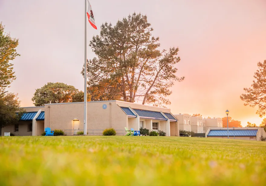 A campus lawn glows at sunset with buildings, trees, and U.S. and a California flag under a sky filled with golden clouds.