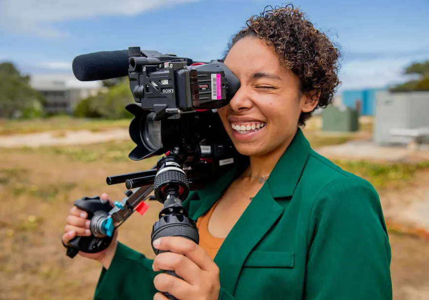 A student films with a professional camera outdoors, capturing creative work in a hands-on media setting.