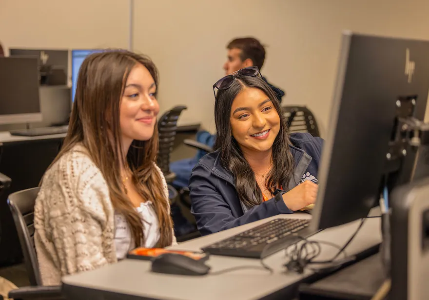 Two girl are smiling and looking atorientation, student collaboration, computer lab, business administration, peer mentoring, campus learning, academic support, new student experience a computer screen