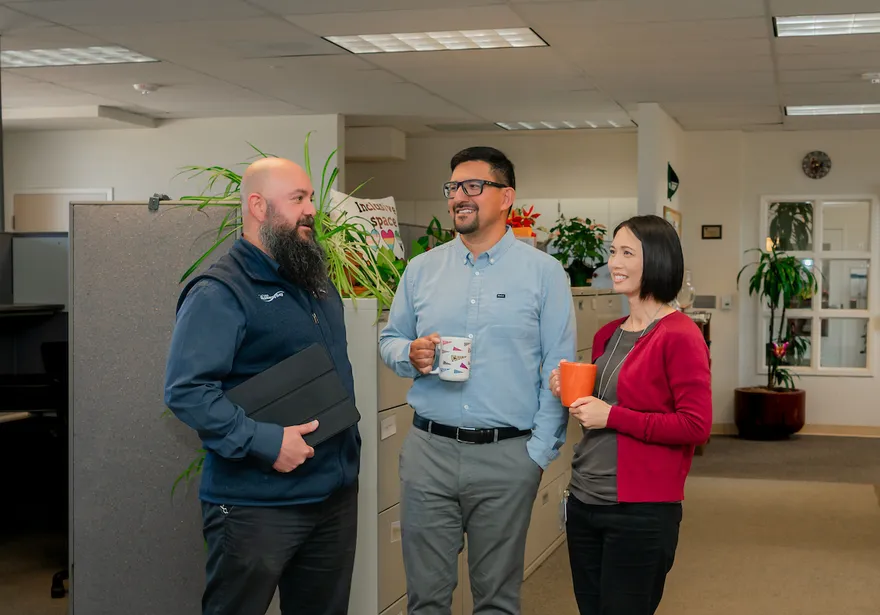 Three ���ӽ紫ý HR staff members share a friendly conversation near a cubicle partition lined with plants and an “Inclusive Space” sign, smiling and holding coffee mugs and a tablet.
