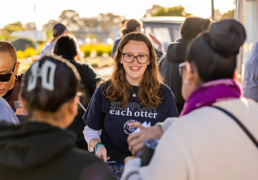A smiling Cal State Monterey Bay staff member wearing a “Support each otter” shirt greets admitted students and families during Admitted Otter Day, engaging in conversation at an outdoor campus welcome event.