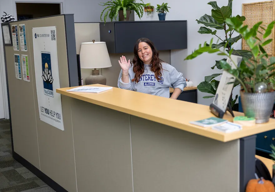 A woman stands behind a desk, smiling warmly and waving hello.