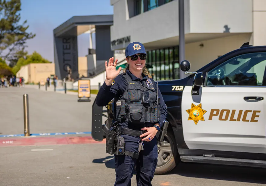 A female CSUMB UPD sergeant waves and smiles at the camera while standing in front of a police SUV on campus.