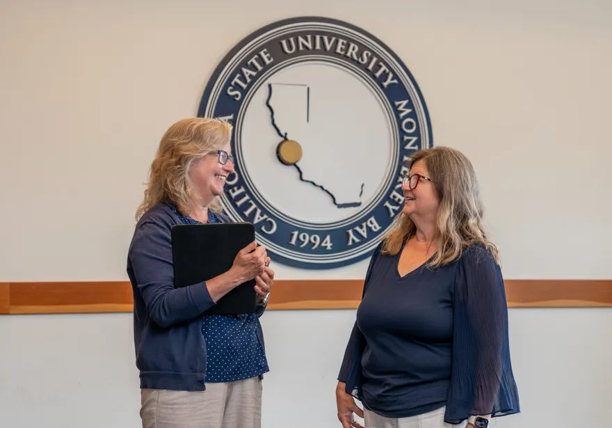 Two staff members chat and smile in front of the California State University Monterey Bay seal inside the Sponsored Programs office.