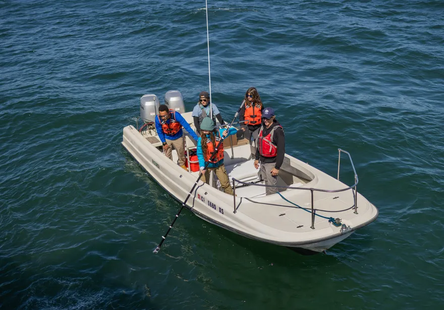 A group of four students and a professor on a boat with life jackets on lowering a pole into the ocean