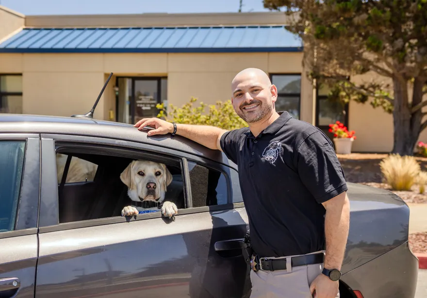 A ���ӽ紫ý officer smiles while standing next to a car window where the service dog looks out with its paws on the door.