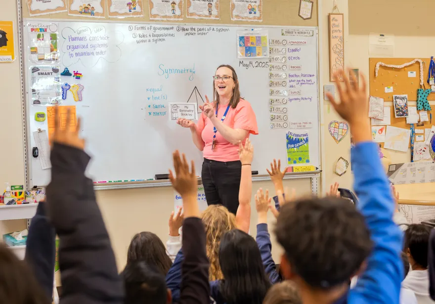 Teacher stands at the whiteboard while students raise their hands in class