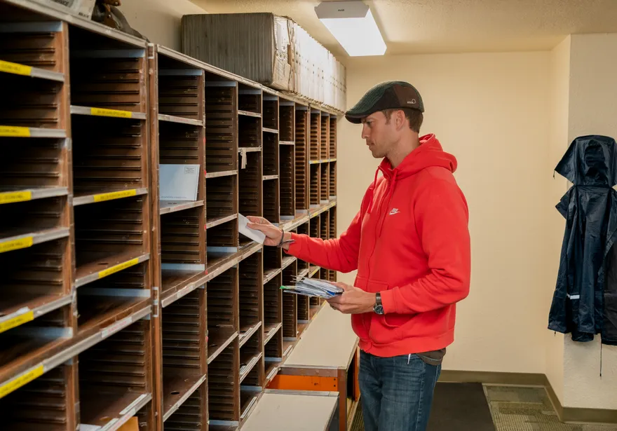 A CSUMB mailroom staff member in a red hoodie sorts mail into a wall of labeled cubbies, ensuring accurate internal campus distribution.