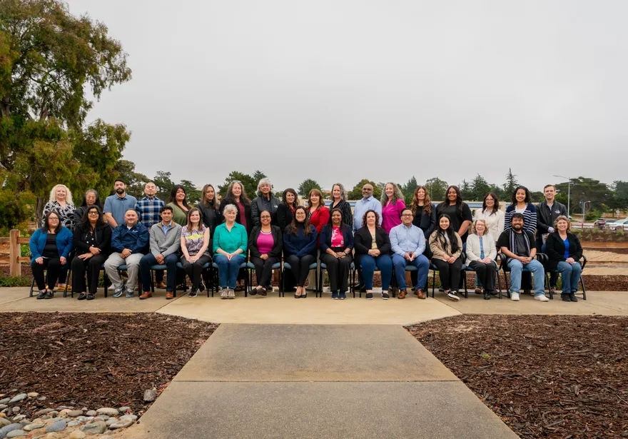 A large group photo of CSUMB’s Finance Department team taken outdoors, featuring staff seated and standing in rows, smiling in front of trees and an overcast sky.