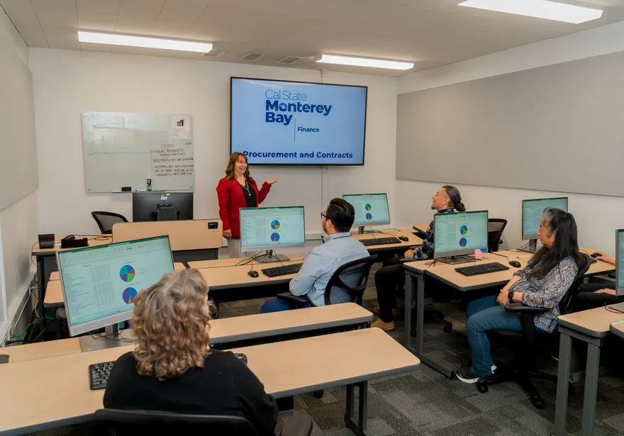 A staff trainer at CSUMB presents a Procurement and Contracts session in a finance department training room as staff listen attentively from their computers.