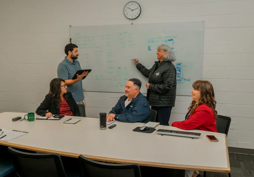 A 911���ϱ����� finance team collaborates during a whiteboard meeting, with one person actively writing notes while others observe and contribute around the table.