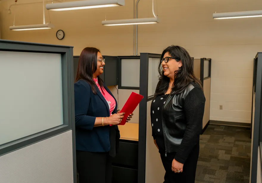 A CSUMB finance staff member holding a red folder shares a conversation with a colleague in the hallway of an office suite lined with cubicles.
