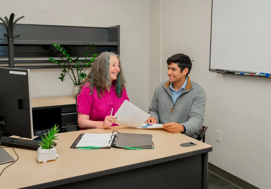 A CSUMB finance team member meets one-on-one with a student in a welcoming office space, smiling and reviewing documents across the desk.